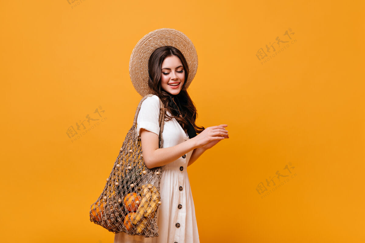 卷发年轻的黑发女孩拎着一个带水果的购物袋戴着帽子和白裙子的女士在橙色的背景下低头女性黑发包