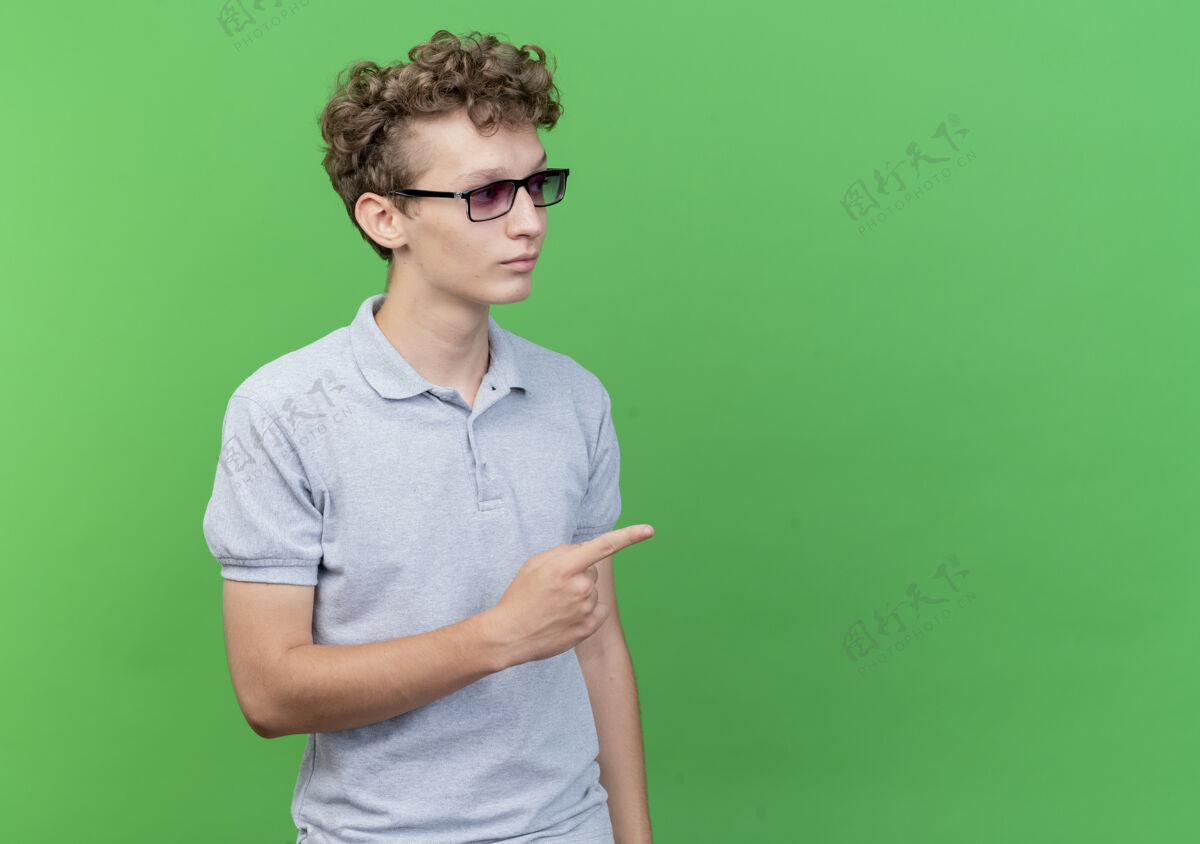 穿一个戴着黑眼镜的年轻人 穿着灰色马球衫 严肃地看着一边 用食指指着绿色的东西年轻指向马球