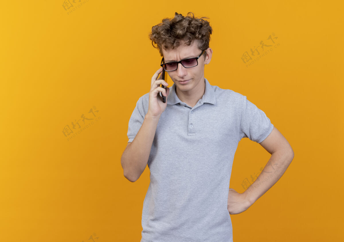 困惑一个戴着黑眼镜 穿着灰色马球衫的年轻人在讲手机 他对橙色感到困惑和不快不高兴手机戴着
