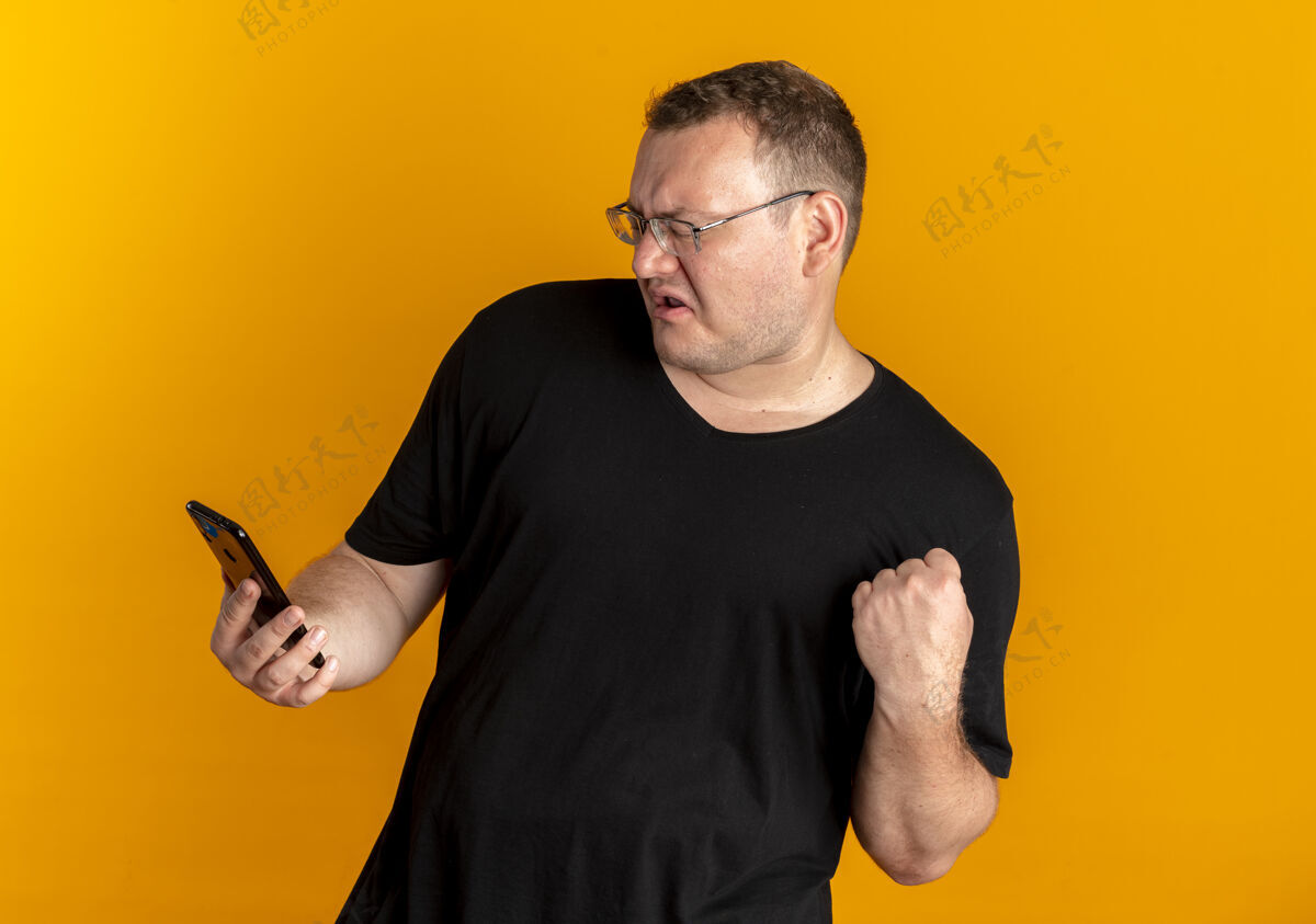 黑色戴眼镜的超重男子身穿黑色t恤手持智能手机握紧拳头 表情咄咄逼人超过橙色站着橙色拳头