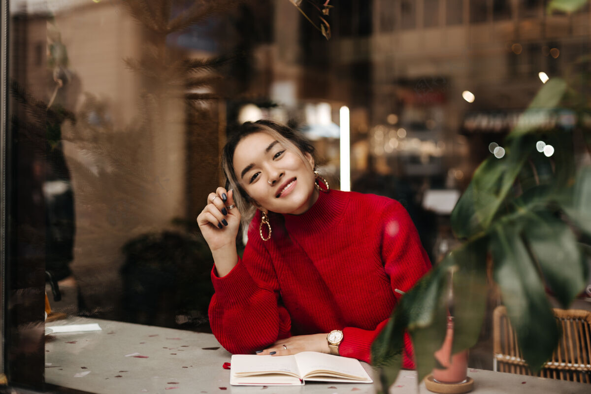 女士迷人的亚洲女人穿着红色针织服装 戴着大大的耳环 坐在咖啡馆里 桌上放着笔记本 微笑着优雅电话华丽