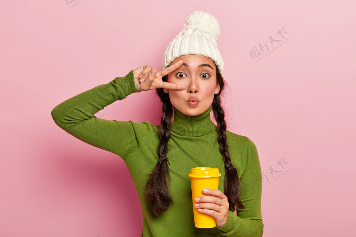 和平年轻的亚洲女孩双唇紧闭 做出和平的姿态 戴着白色暖冬帽 绿色套头衫 喝着咖啡去 靠着粉色的墙站着面部帽子亚洲人