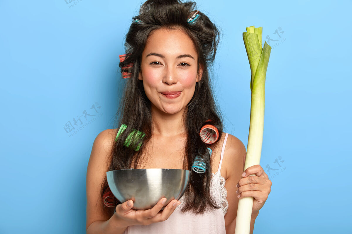 碗令人愉快的满意的亚洲女人 深色头发 拿着钢碗和新鲜蔬菜 做美味的沙拉 留着卷发的深色长发 穿着睡衣 在室内摆姿势请黑发素食主义者