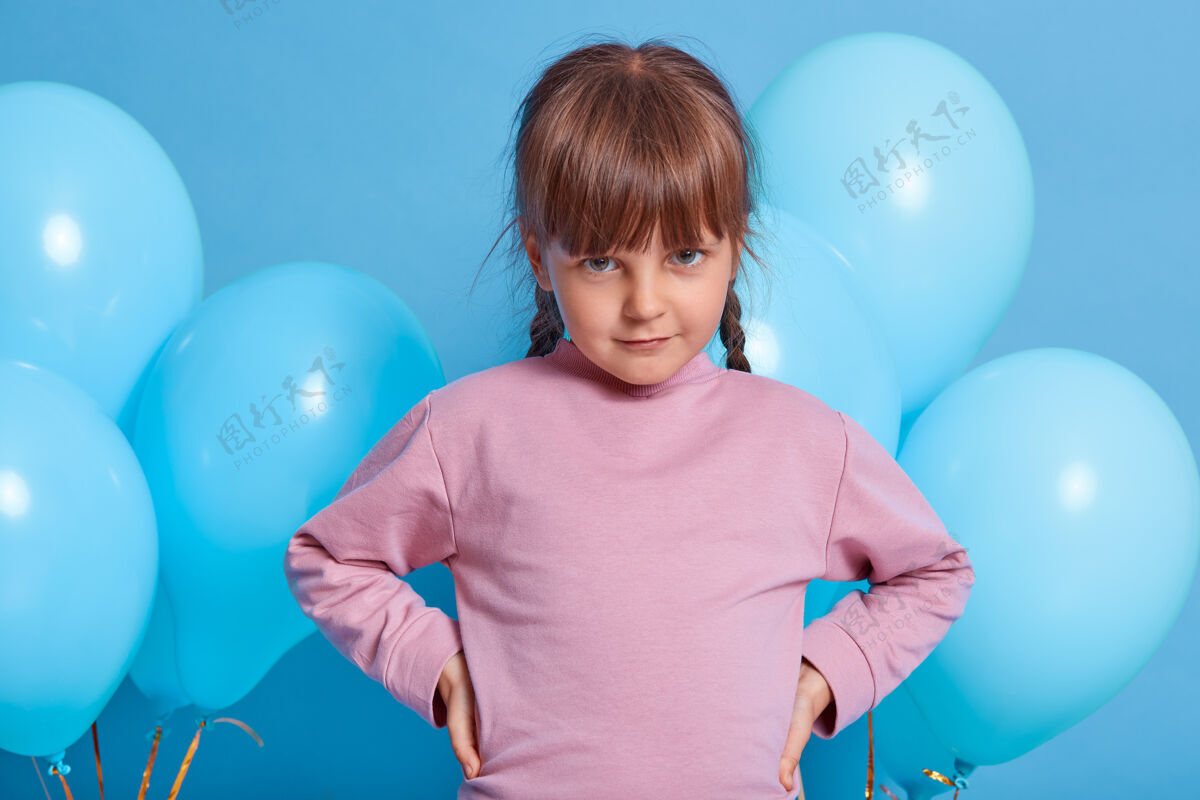 女性腼腆可爱的小女孩和蓝色气球在彩色背景下单独摆姿势漂亮的孩子从额头下看着相机 手放在臀部 穿着玫瑰毛衣欢呼聚会肖像