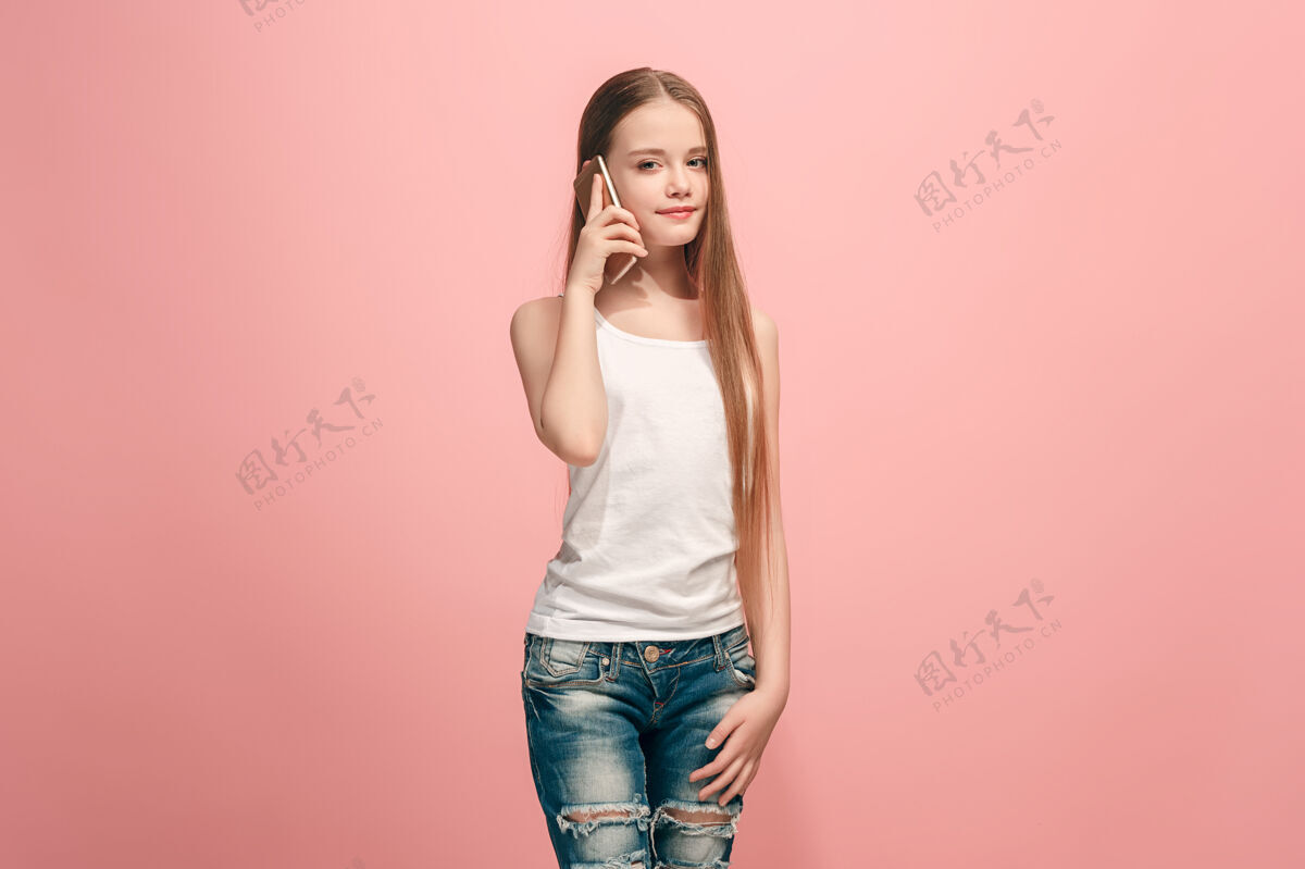 女士快乐的少女站着 微笑着用手机在时尚的粉红色工作室情感青少年女性