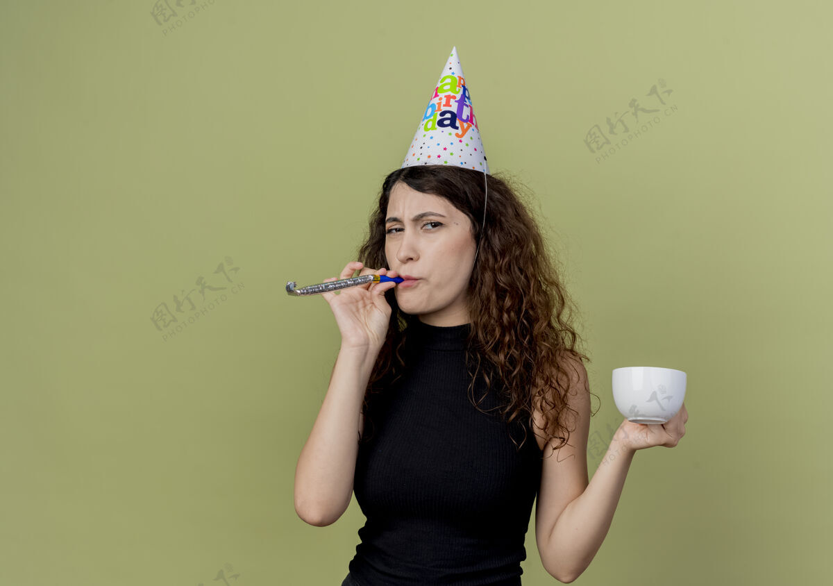 节日年轻漂亮的女人 卷发 戴着节日帽吹着口哨 手里拿着咖啡杯 看起来很不高兴 生日派对的概念在灯光下杯子握着漂亮