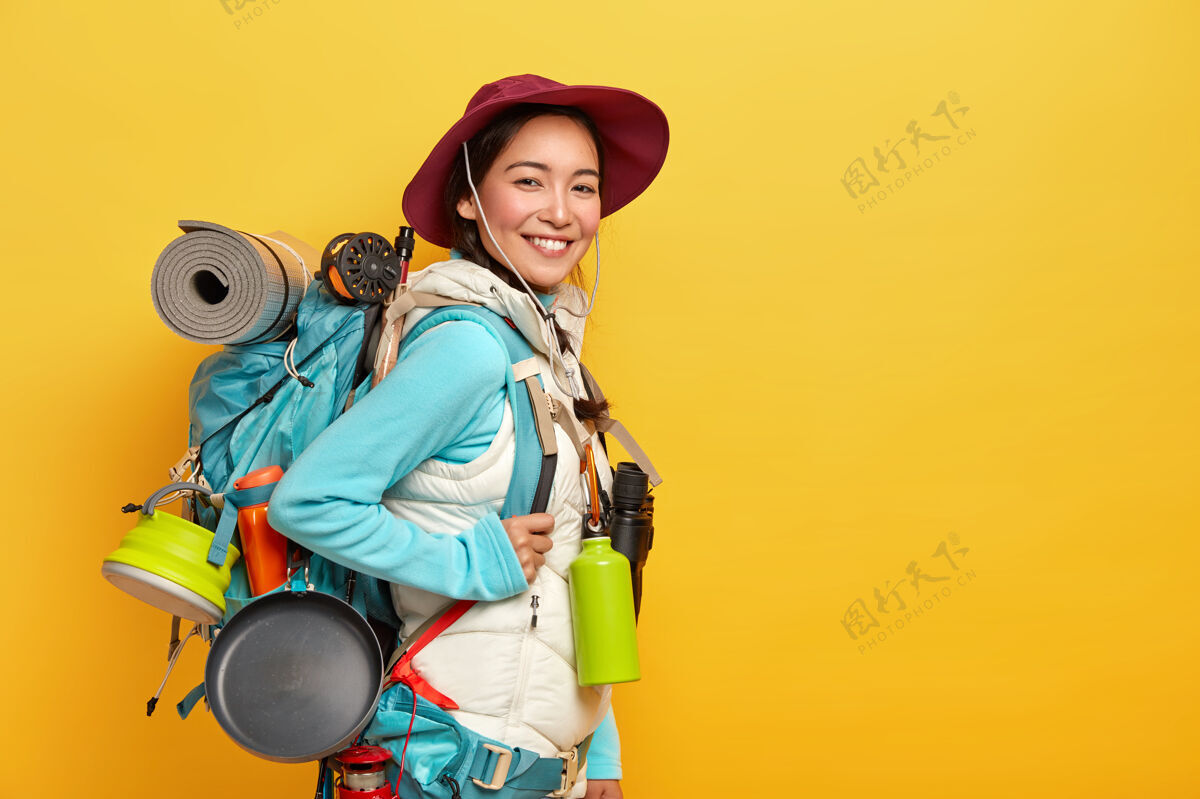 徒步旅行快乐的深色头发的亚洲旅行者背着大大的旅游背包 用望远镜旅行 靠着黄色的墙 戴着时髦的帽子 穿着背心的套头衫帽子看方向