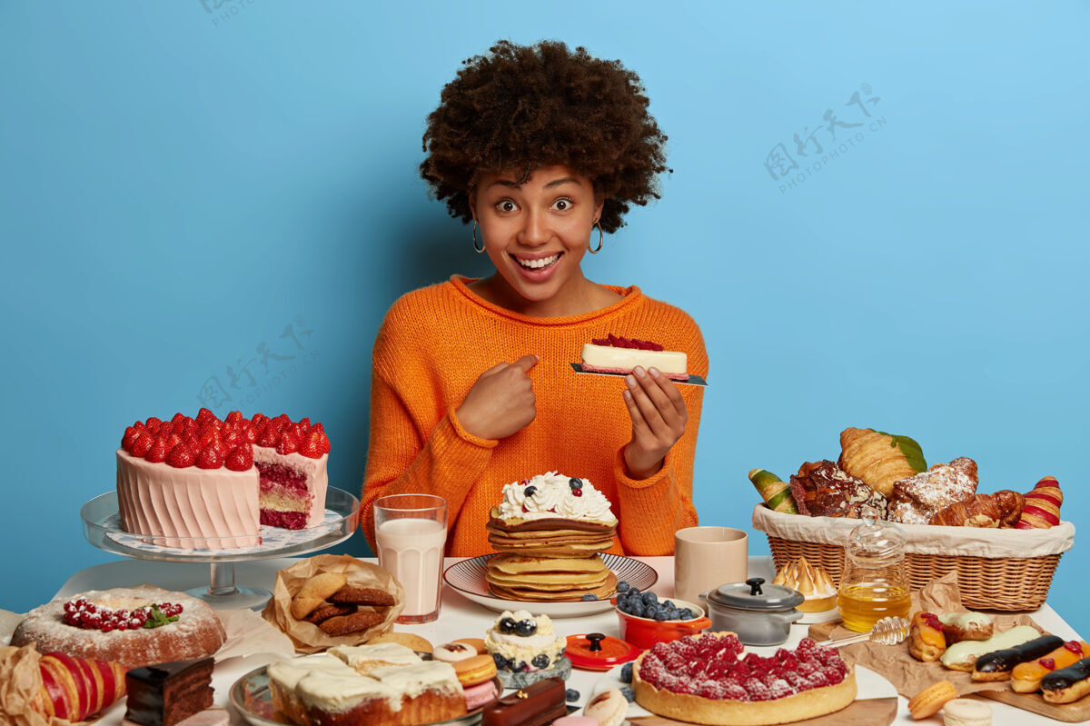 桌子高兴的黑皮肤女人有积极的目光 指着自己 拿着美味的蛋糕 问她是否应该吃它的一切 穿着橙色套头衫 隔离在蓝色的墙上水平盘子满意