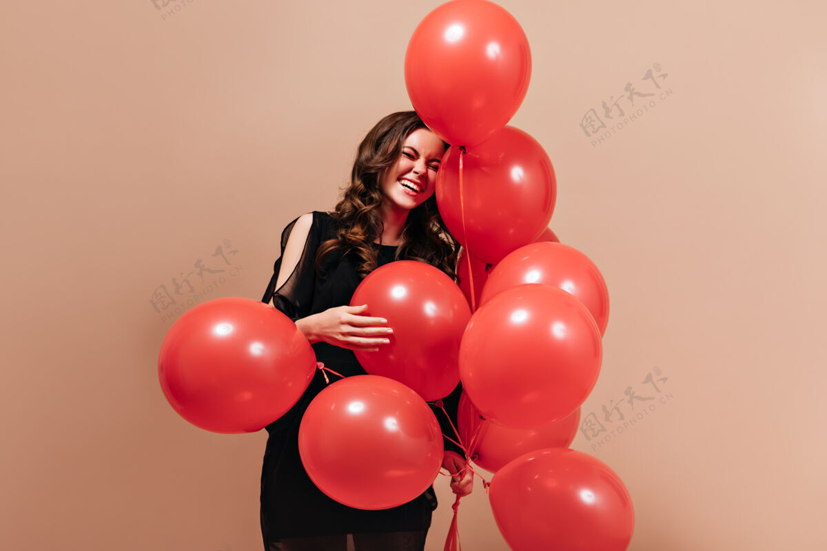 优雅在孤立的背景下 穿着黑色套装的积极的卷发女人笑着和红气球摆姿势派对黑发女性