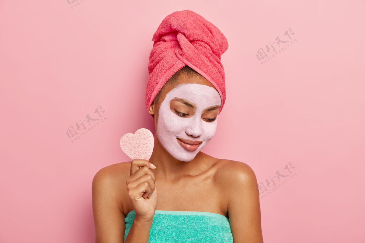 治疗高兴的年轻女子专注下来 应用粘土面膜 举行化妆海绵卸妆 显示裸露的肩膀 裹在浴巾 隔离在粉红色的工作室墙上清洁皮肤护理满意