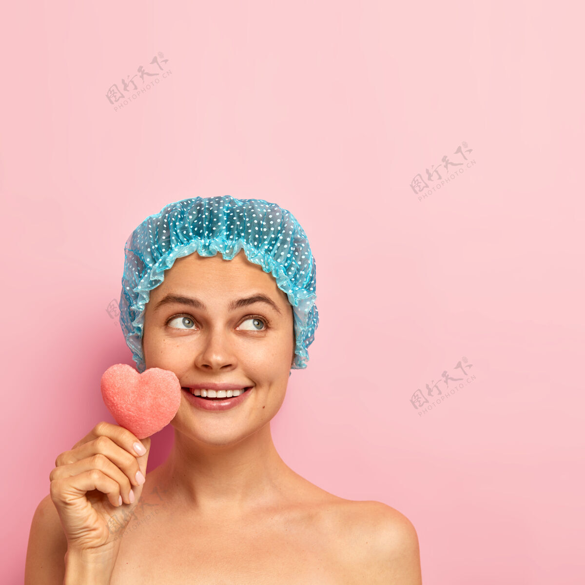 护理快乐快乐的年轻女子有着精心呵护的皮肤 牙齿般的微笑 神态如梦的站着 拿着小化妆海绵 戴着保护性的淋浴帽化妆面部模特