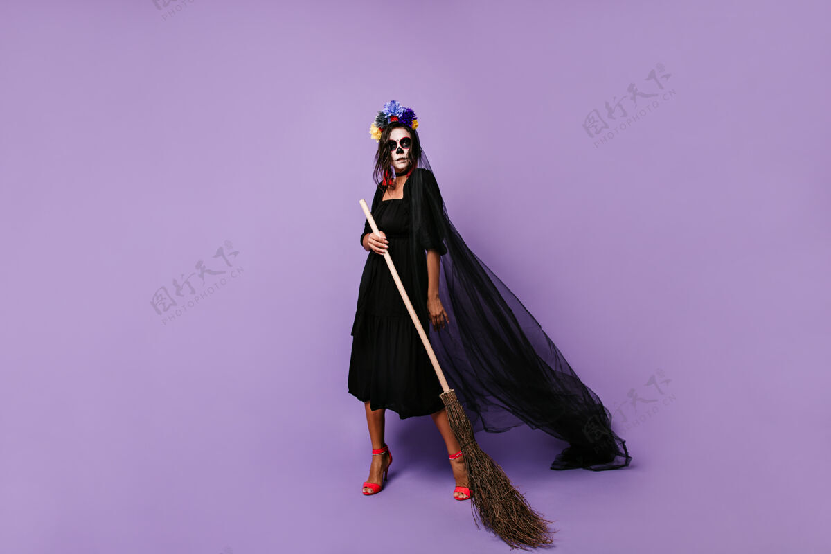 恐怖女巫戴着骷髅面具 穿着黑色的令人毛骨悚然的服装女人拿着扫帚在淡紫色的墙上摆姿势墨西哥女孩墨西哥