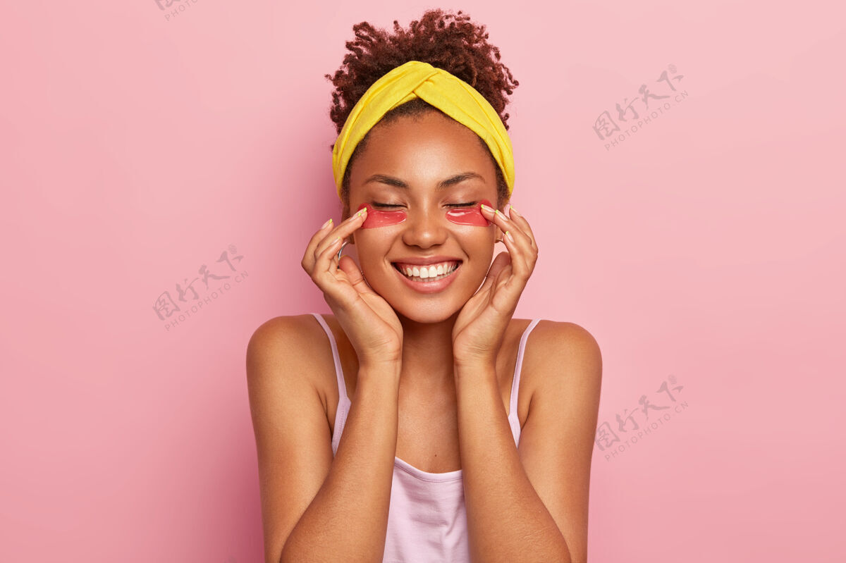 眼睛年轻的非洲裔女模特在眼睛下面涂上胶原蛋白护垫 享受保湿治疗 笑容灿烂 露出洁白的牙齿 有新鲜健康的皮肤 戴着黄色的头巾恢复水凝胶健康