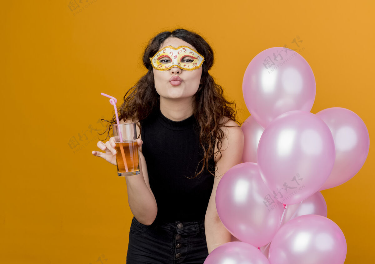 卷发年轻漂亮的卷发女子手持一束气球和鸡尾酒 戴着派对面具 开心地吹着生日派对的飞吻站在橘色的墙上面具女人吹