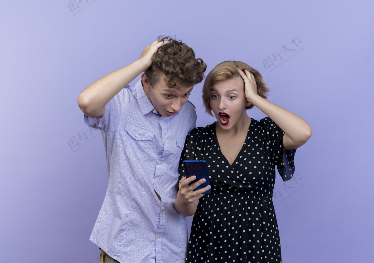 屏幕年轻漂亮的情侣们站在蓝色的墙上 看着智能手机的屏幕惊讶又惊奇女人手机看