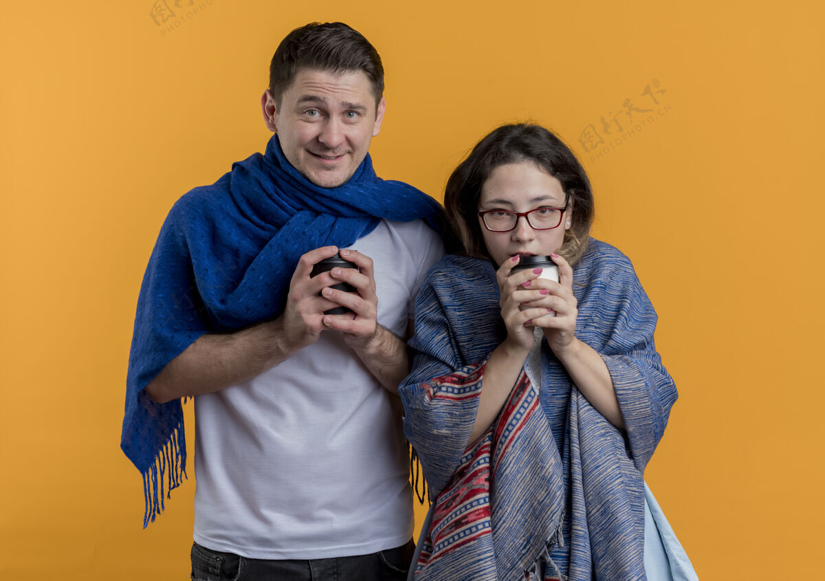 拿着冻住的年轻夫妇拿着毯子拿着咖啡杯站在橙色的墙上冷冻的杯子毯子