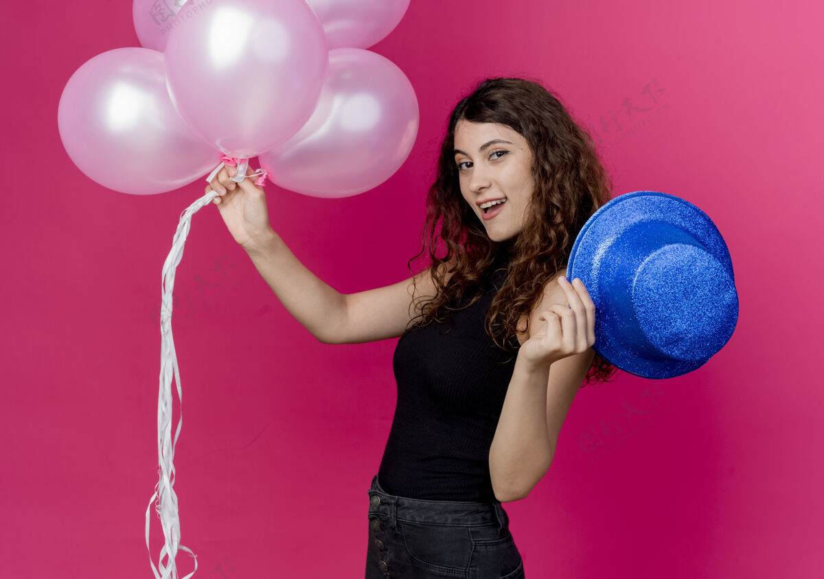 微笑一位年轻漂亮的女士 卷发 手持一束气球 戴着节日礼帽 站在粉红色的墙上 开心地微笑着束空气年轻