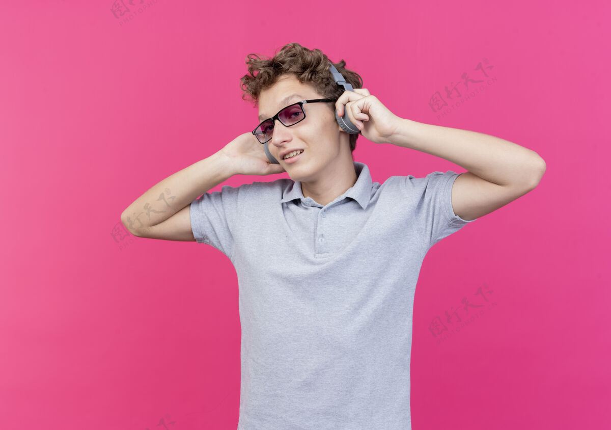 年轻戴着黑眼镜的年轻人 穿着灰色马球衫 戴着耳机 站在粉色的墙上愉快地享受着最喜爱的音乐享受微笑站
