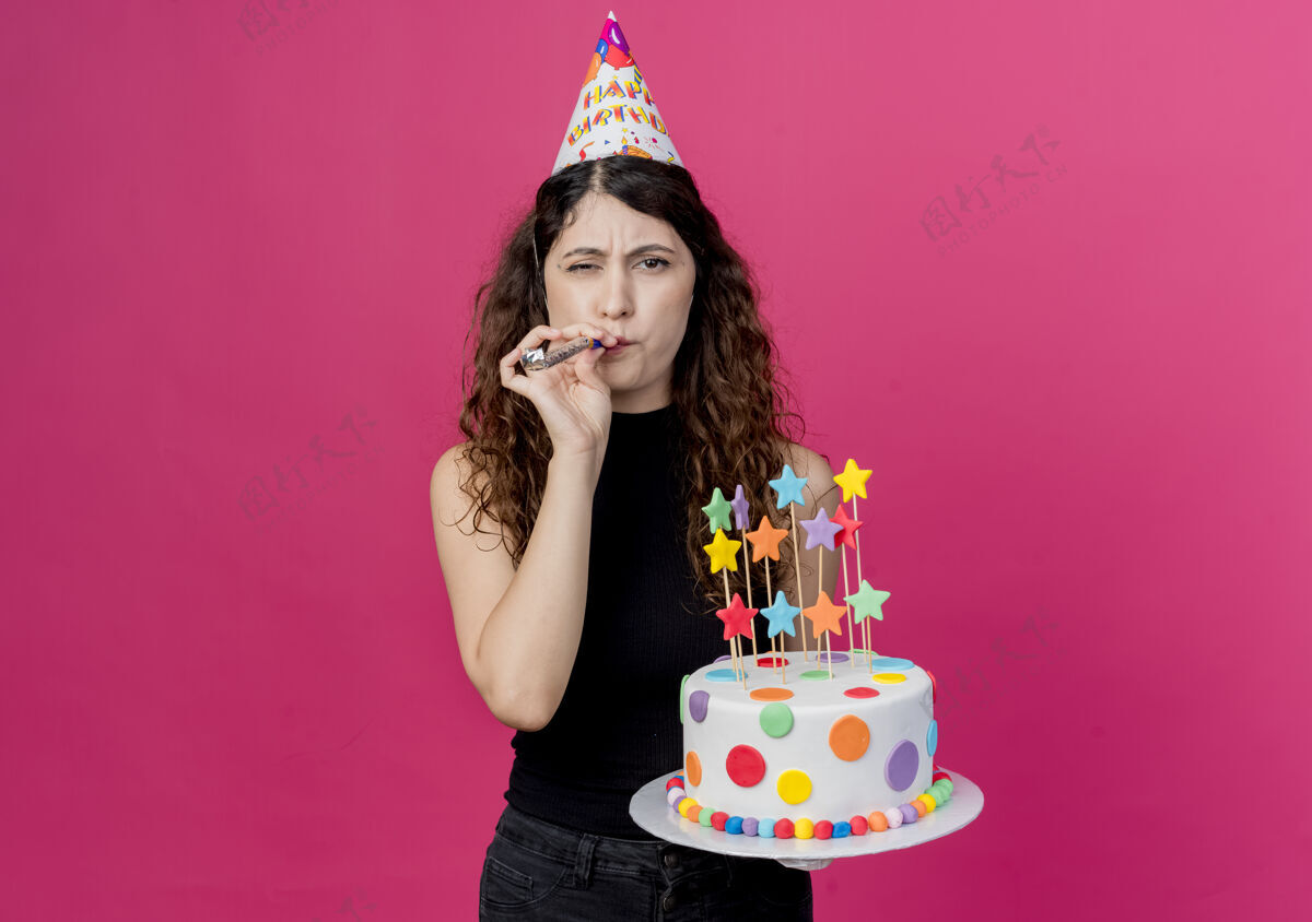 站着年轻漂亮的女人卷发戴着节日帽拿着生日蛋糕吹着口哨站在粉色的墙上快乐而积极的生日派对理念年轻积极向上抱着