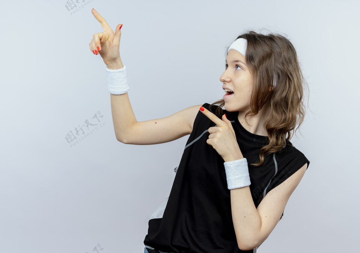 手指身穿黑色运动服的年轻健身女孩戴着头带微笑 手指指向站在白墙上的一边微笑壁板健身