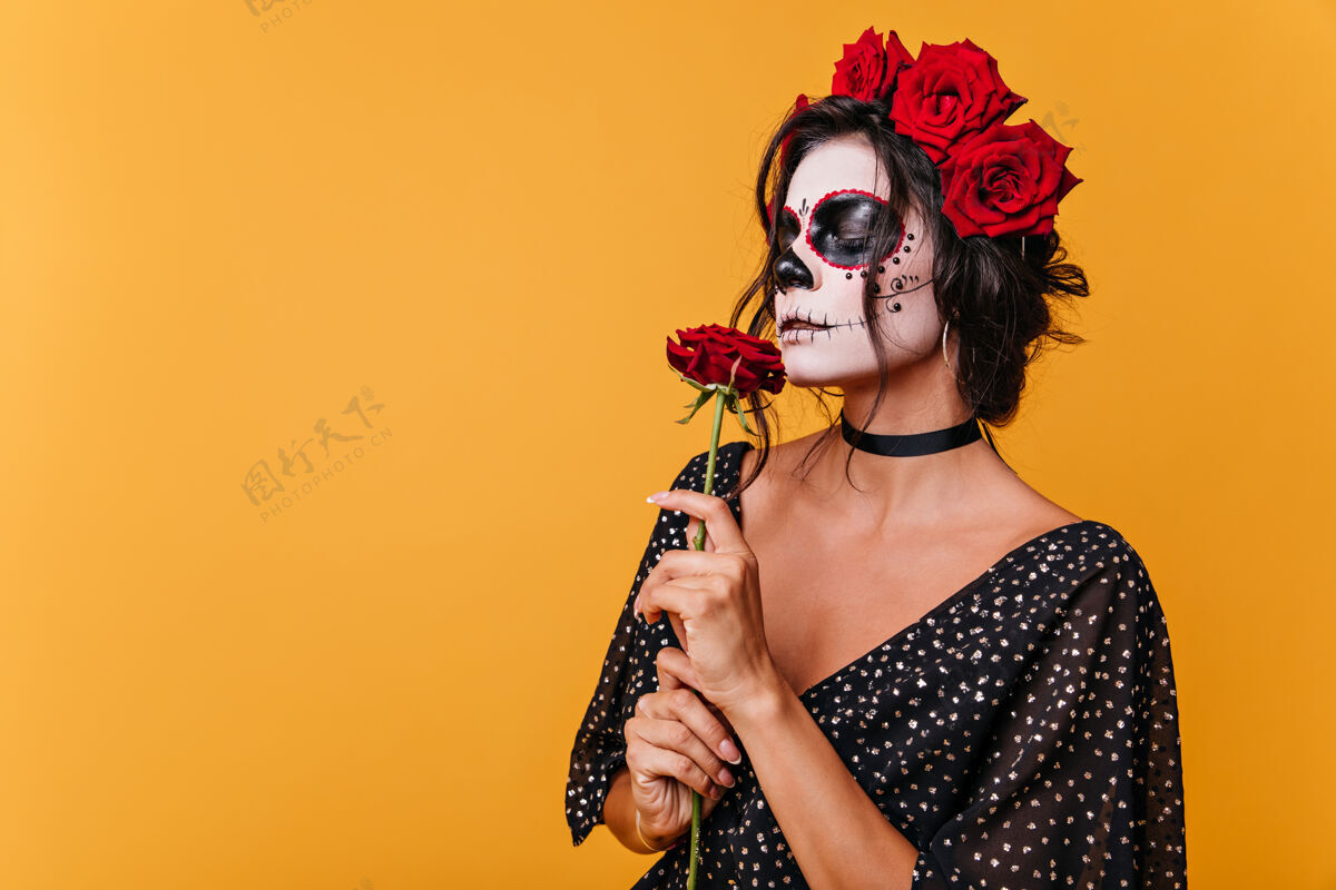 魔鬼万圣节戴着僵尸面具的女孩闻到了芬芳的玫瑰味橙色墙上穿着黑色连衣裙的模特肖像恐怖可怕