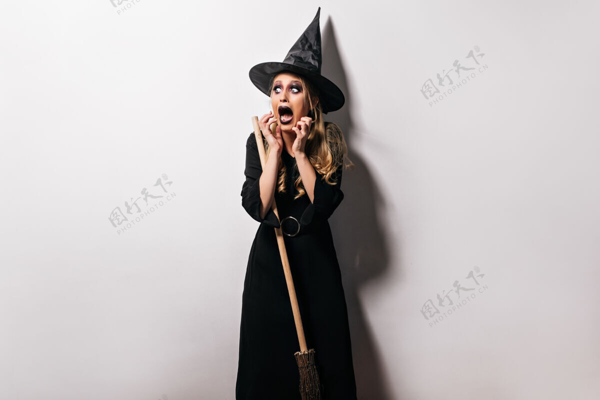 魔术戴着帽子的小女巫在万圣节摆出一副吓人的表情穿着巫师服装的金发美女模特的室内照片女巫邪恶万圣节