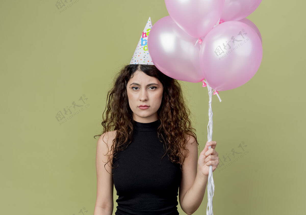 帽子戴着节日帽 卷发的年轻漂亮女人举着皱着眉头的气球站在光墙上生日派对的概念脸年轻看