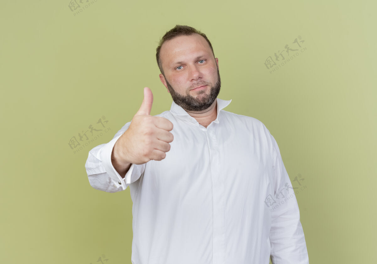 显示一个留着胡子的男人穿着白衬衫 竖起大拇指 微笑着 自信地站在明亮的墙上光穿男人