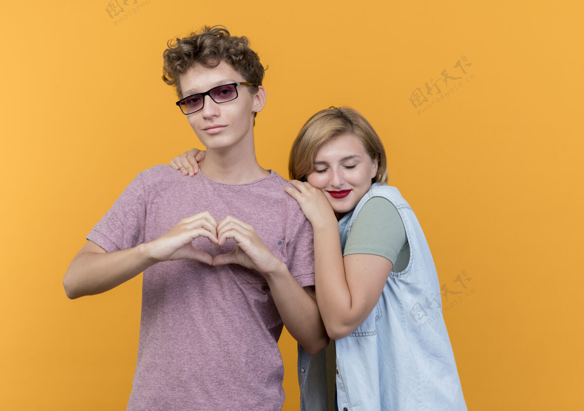 肩膀年轻漂亮的一对男女穿着休闲服站在一起 男人在做手势 他的朋友把头靠在他的肩膀上 站在橙色的墙上而穿衣服