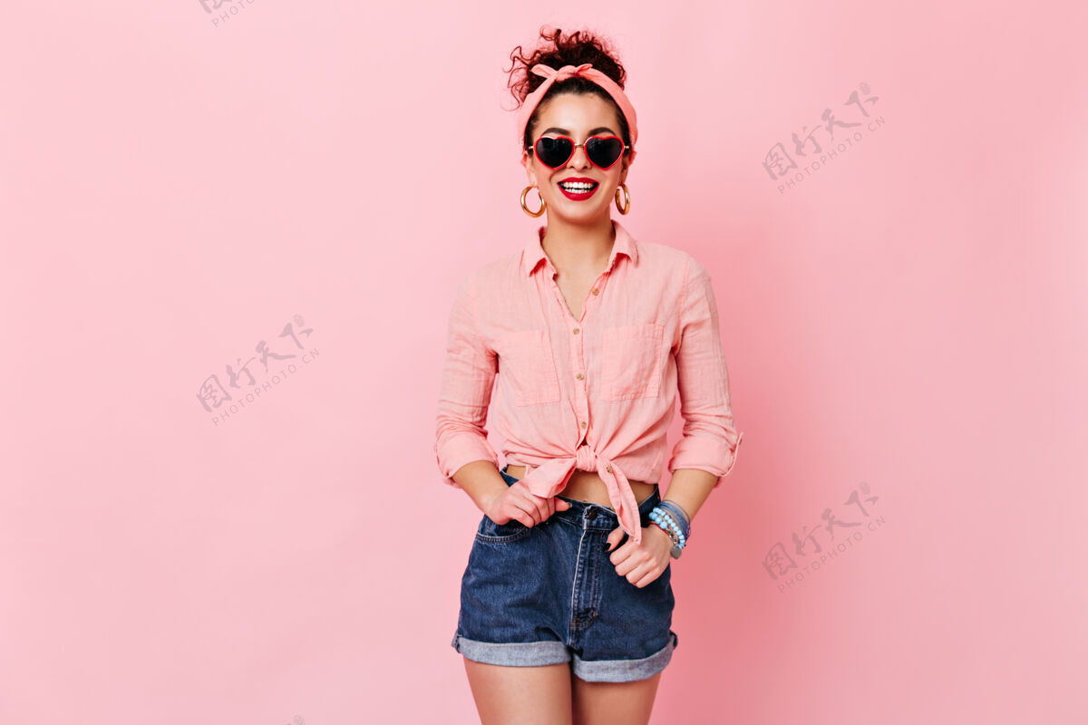 调情心情很好的女人在粉红色的空间里摆姿势戴着墨镜和金耳环 穿着衬衫和短裤的女孩在微笑发型太阳镜DIVA