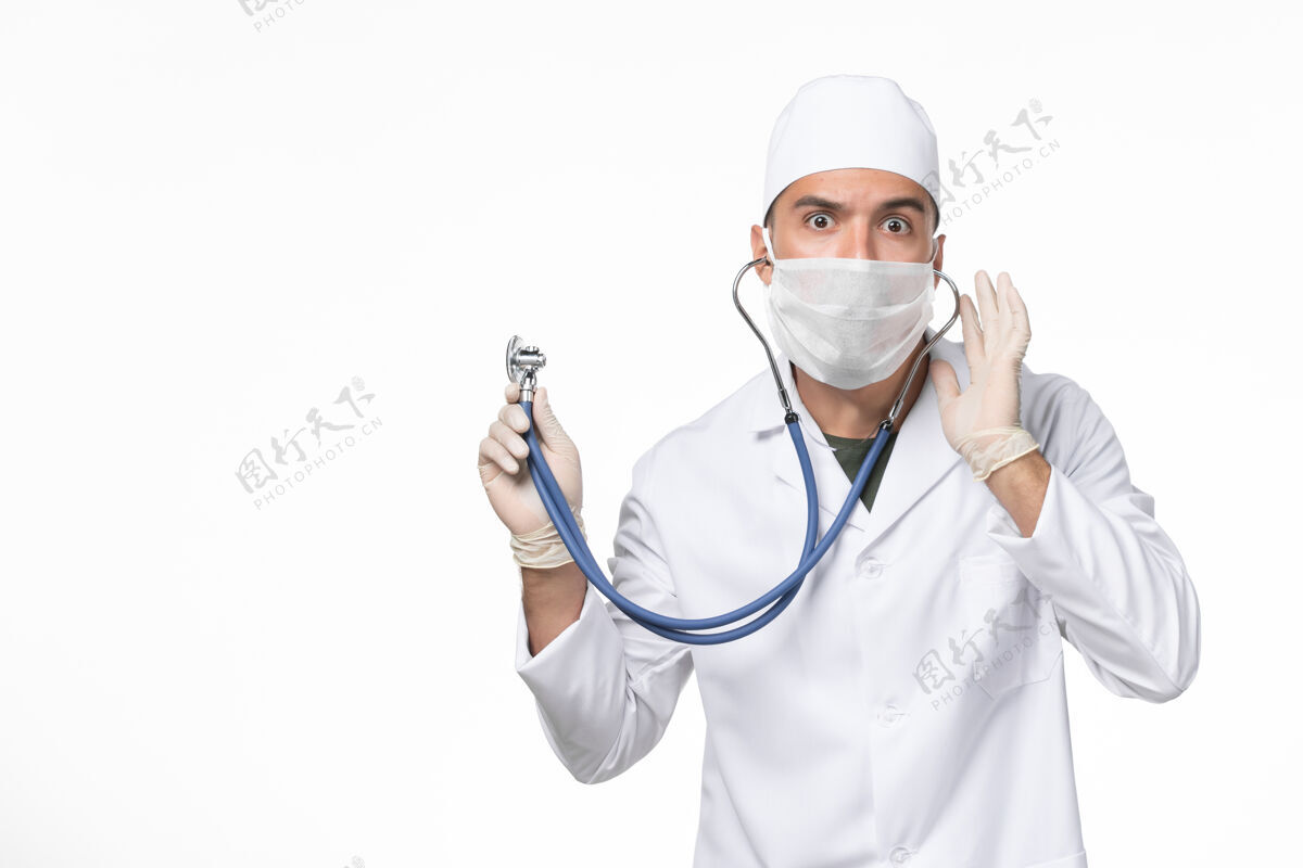专业正面图：男医生穿着医疗服 戴着口罩 由于冠状病毒-使用听诊器对白色墙壁冠状病毒-大流行疾病冠状病毒药品冠状病毒预防