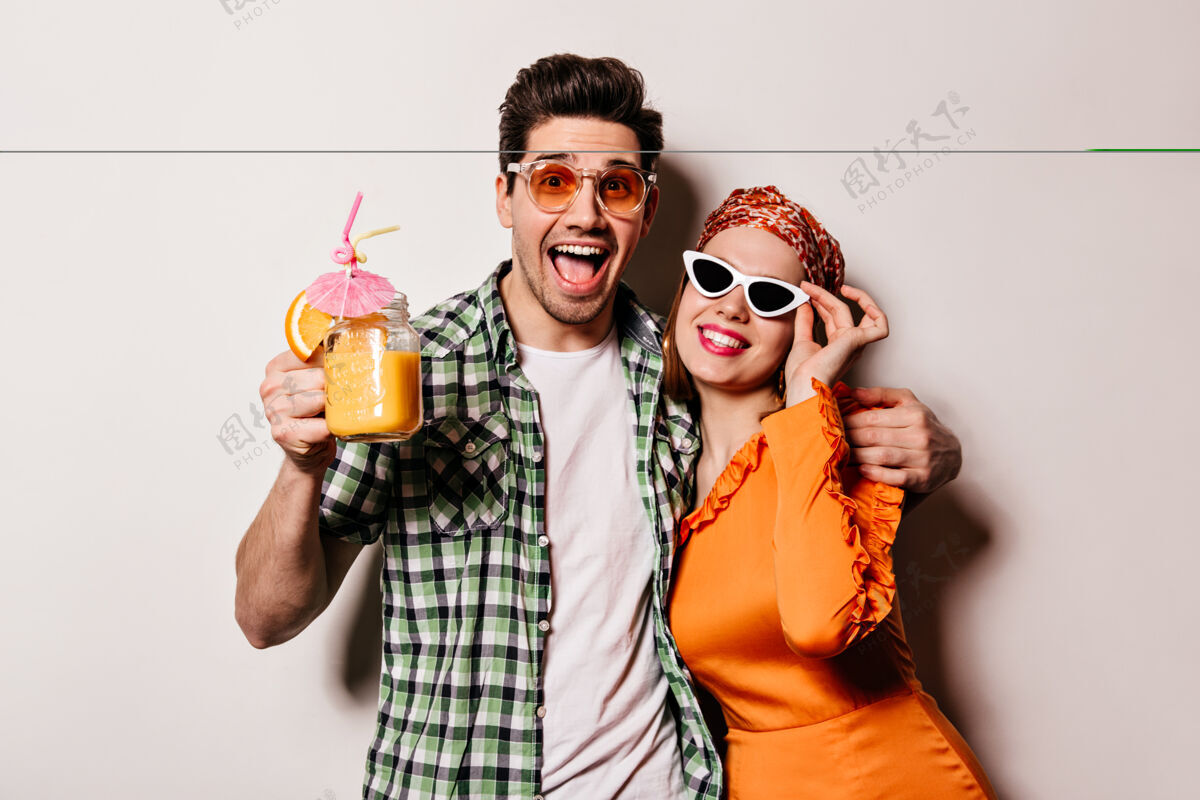 手淘气的男孩和女孩在时尚的服装和太阳镜拥抱 微笑和摆姿势与橙色鸡尾酒在白色空间老式相机情侣男性