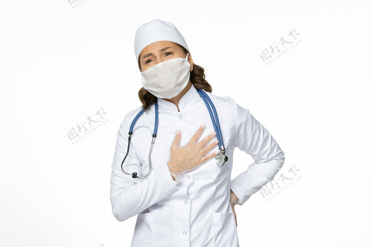 医疗白壁上有心痛的身穿白色医疗服和口罩的女医生pandemy病毒病药专业听诊器疾病