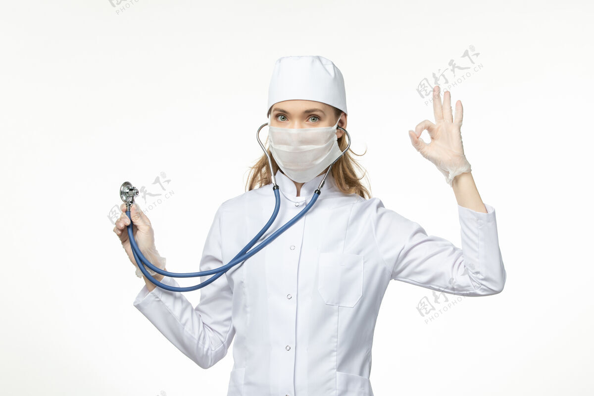 疾病正面图女医生穿着白色医疗服 带无菌口罩 因冠状病毒使用听诊器对白墙疾病冠状病毒-大流行性疾病正面视图大流行
