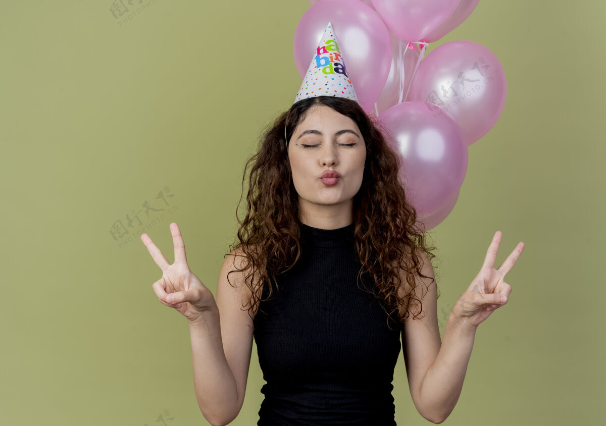 女人一位年轻漂亮的女士 卷发 戴着节日帽 快乐而积极 闭着眼睛站着 手里拿着气球站在光墙上光立场关闭