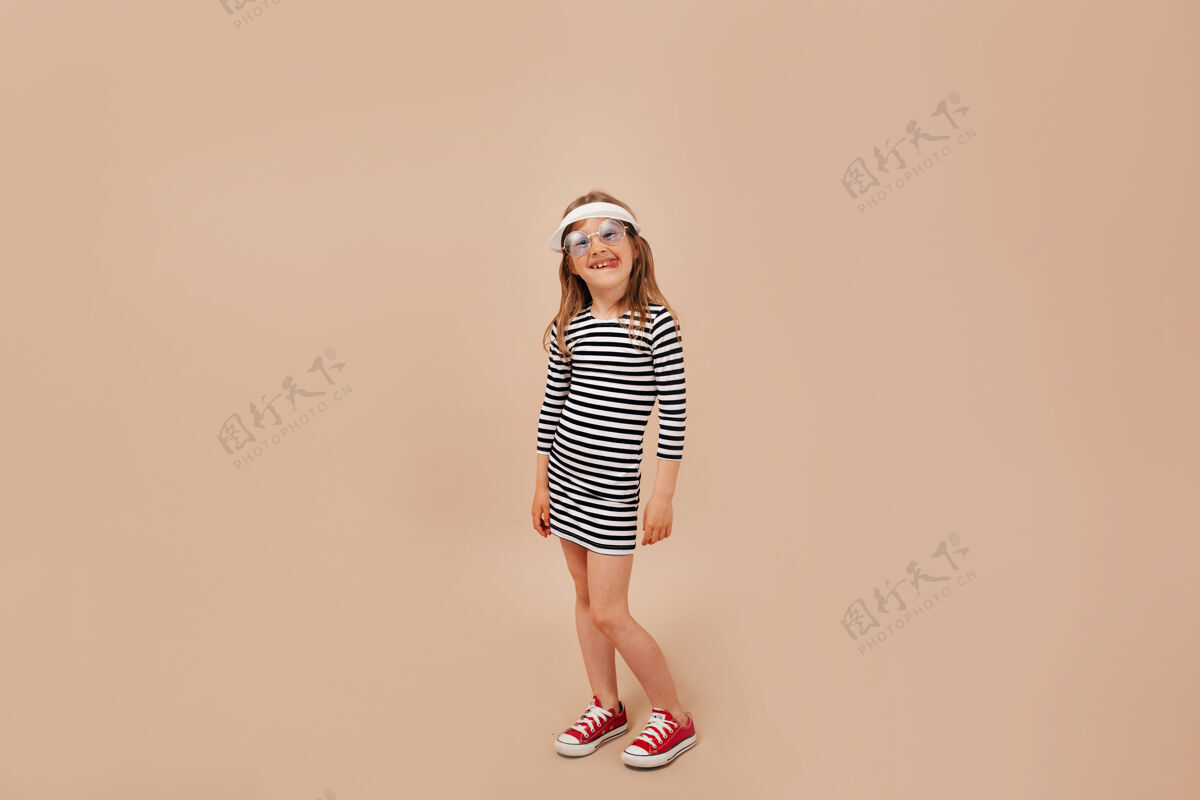 孩子全长度的照片可爱迷人的小女孩穿着裙子 帽子和运动鞋摆在相机在米色背景微笑幼儿全长