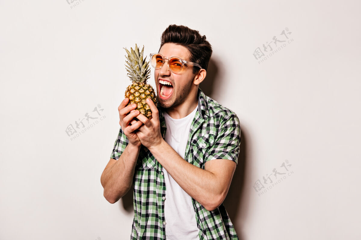 棕色眼睛一个穿着白色t恤 绿色衬衫 戴着明亮太阳镜的开朗的家伙想咬下一片菠萝深色水果肌肉