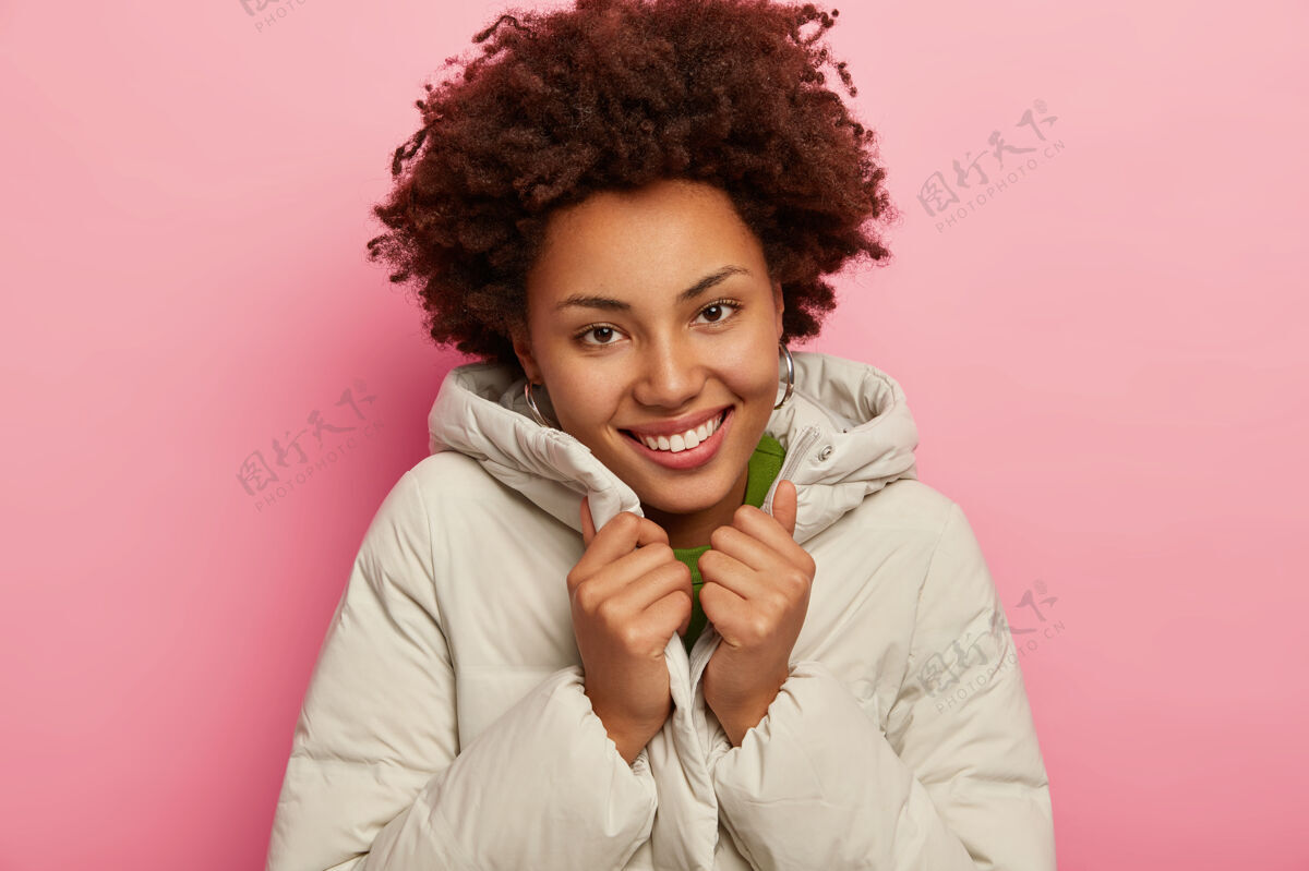 罗西美丽的快乐女人 卷发 穿着温暖舒适的外套 健康的深色皮肤 微笑愉快 与玫瑰色背景隔离请情感牙齿