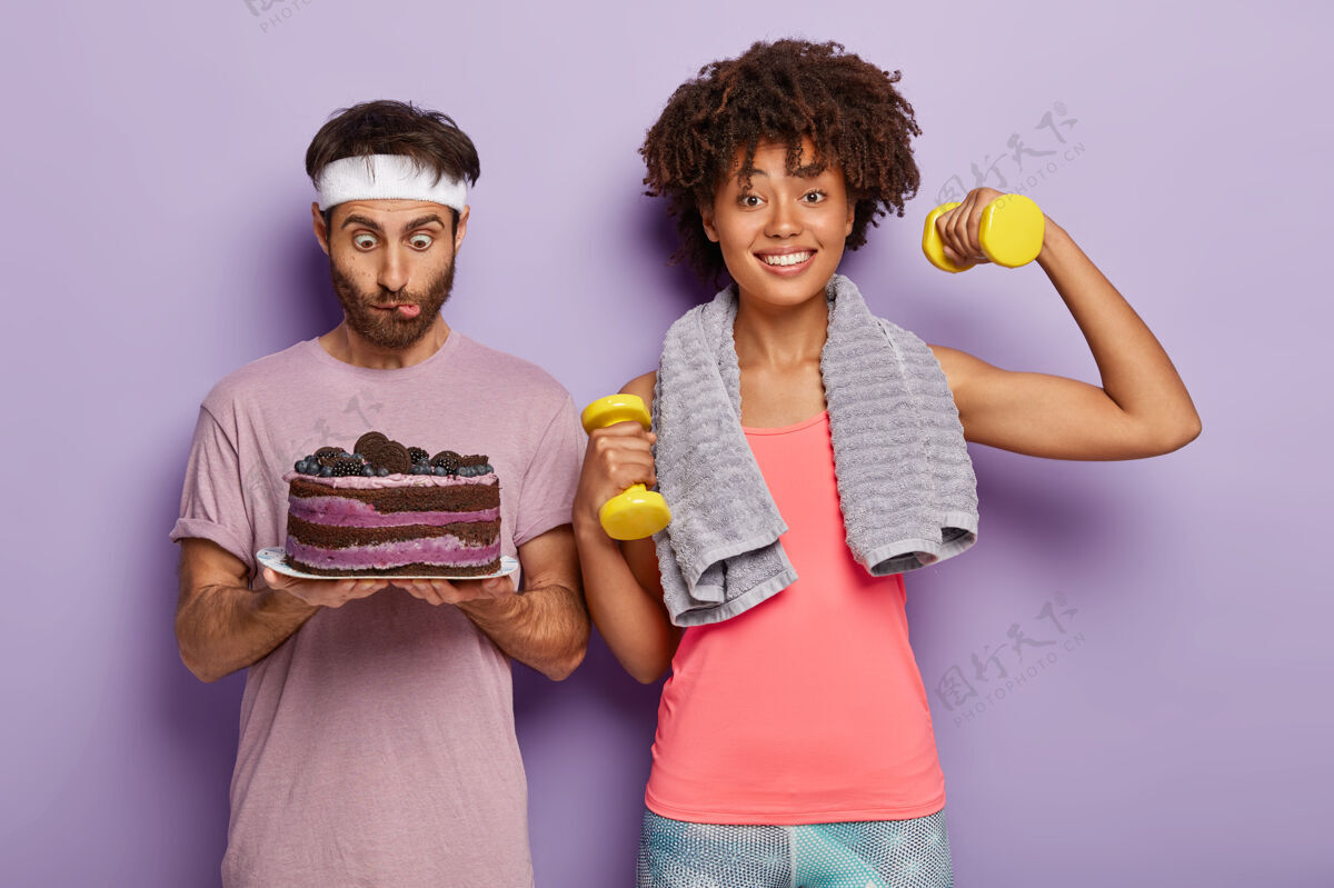 紫色惊讶的年轻非洲男人盯着美味的蛋糕 戴着白色的头带 感受着诱惑 快乐的女人锻炼二头肌 举重 过着运动的生活 站在紫色的背景下诱惑震惊蛋糕