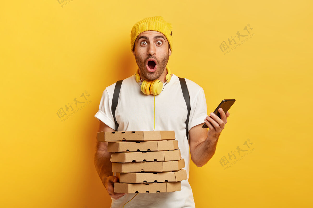 男性惊奇送货员通过智能手机接收顾客的订单 拿着一堆纸板比萨饼盒 背着背包 戴着帽子和t恤 隔离在黄色背景下 在餐厅工作快递食物堆