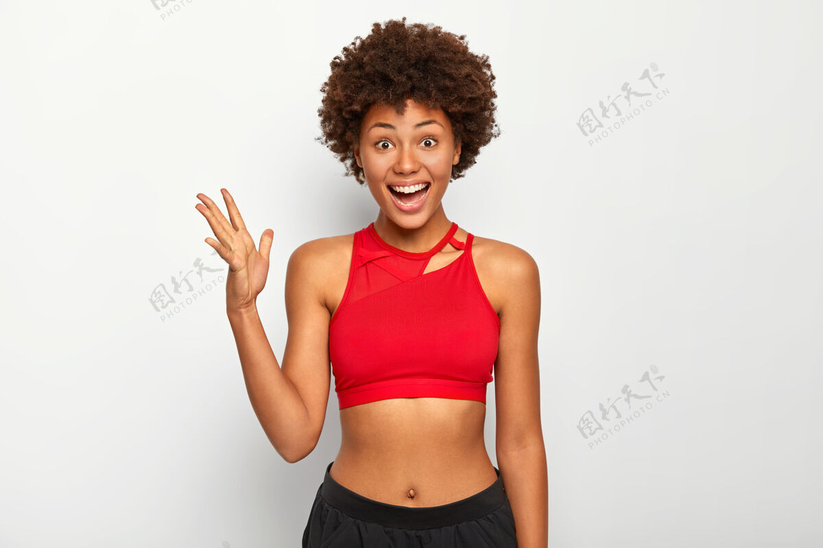 运动这张照片是一位开朗的黑皮肤女性 她举着手掌 穿着红色运动胸罩 露出腹部 留着非洲式发型 在白色背景下在室内摆姿势手美国黑