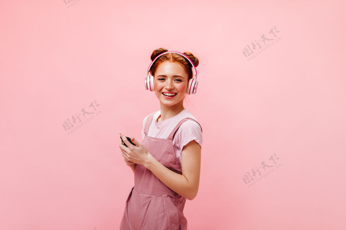 女性带着发髻的年轻女士惊讶地看着手机屏幕穿着裙子和白色t恤的女士正在用粉色背景的耳机听音乐成人肖像优雅