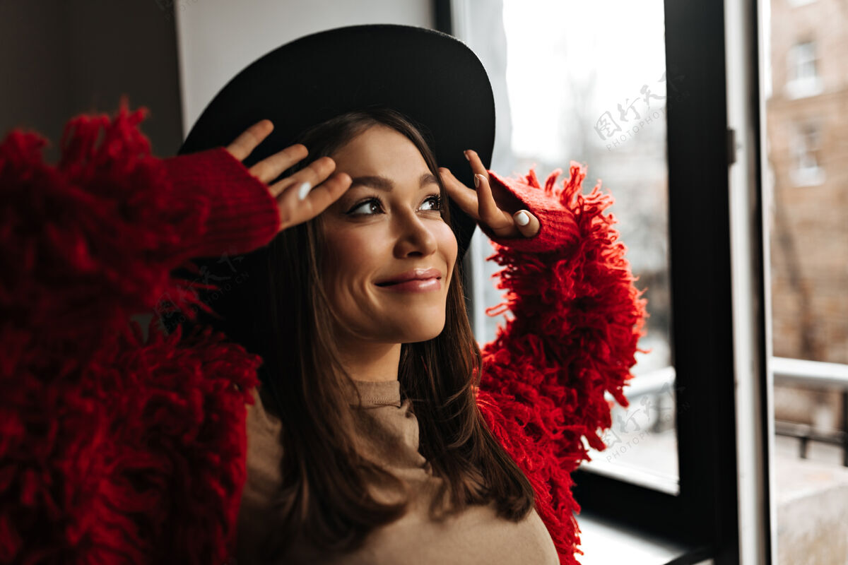 美丽乐观的女士在心情好的时候涂上唇彩 看着窗外穿着红色环保外套和帽子的女人的照片魅力羊毛漂亮