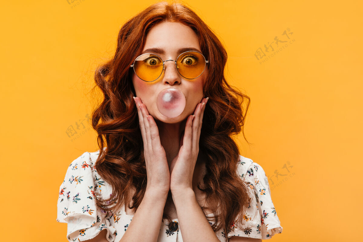 年轻戴着黄色眼镜的淘气女人在橙色背景上吹口香糖泡肖像美丽包