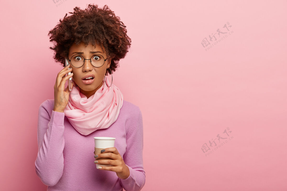 麻烦不开心 紧张 震惊的非裔美国妇女通过手机交谈 拿着外卖咖啡 听到坏消息 戴着眼镜和紫色的马球衫 在玫瑰色的工作室墙上摆姿势女人眼镜罗西