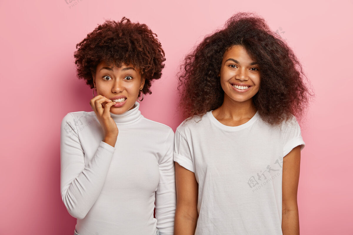 紧张迷惑不解紧张的非裔美国妇女咬指甲 看着焦急的镜头 她兴高采烈的女朋友披着浓密的卷发站在旁边 穿着白色的衣服 表达着不同的情感微笑粉色团友