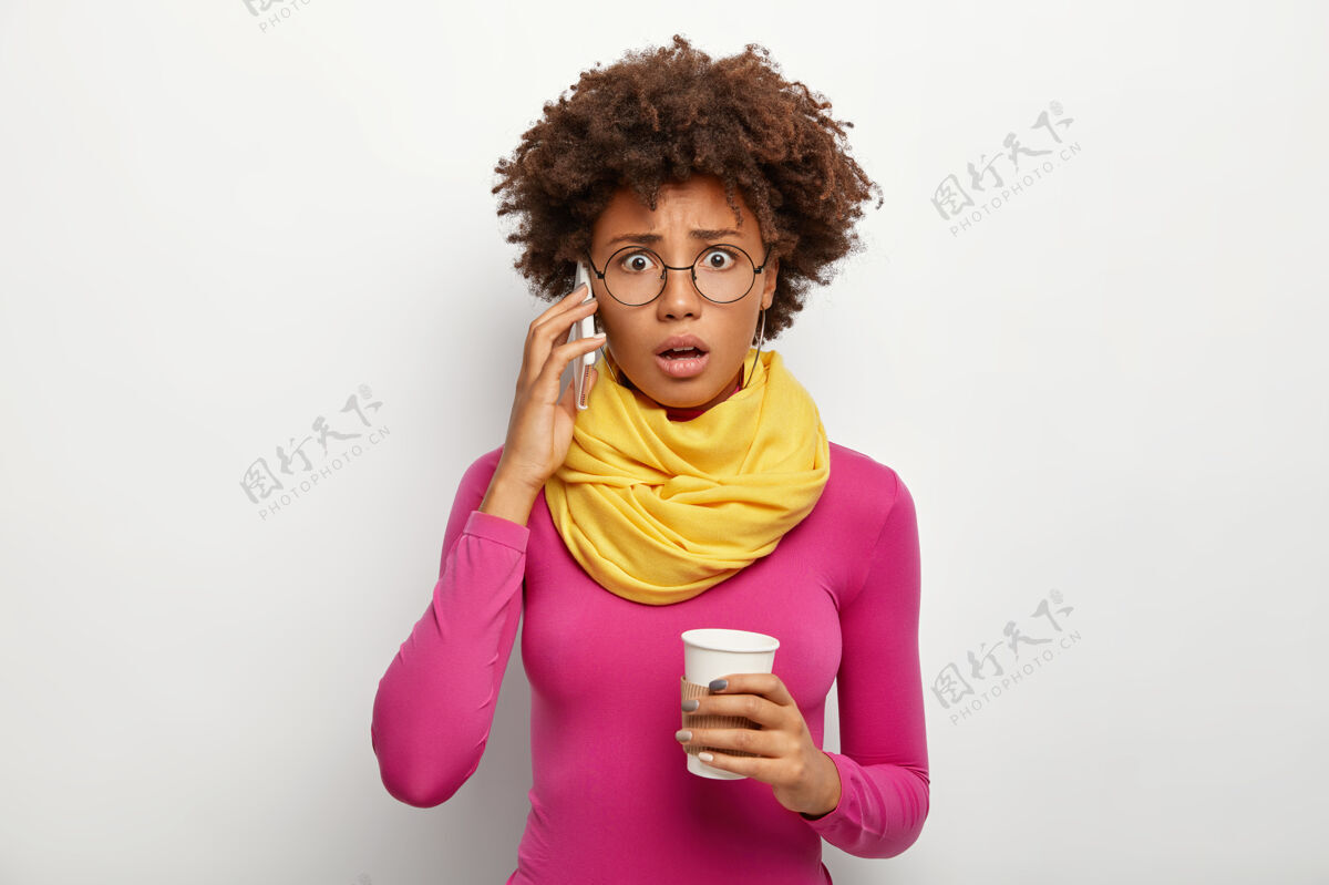 水平尴尬的卷发女人通过智能手机说话 听到不好的信息 打电话 拿着外卖咖啡 透过光学眼镜 圆领衫和围巾看 隔离在白色背景下马球衫非洲眼镜