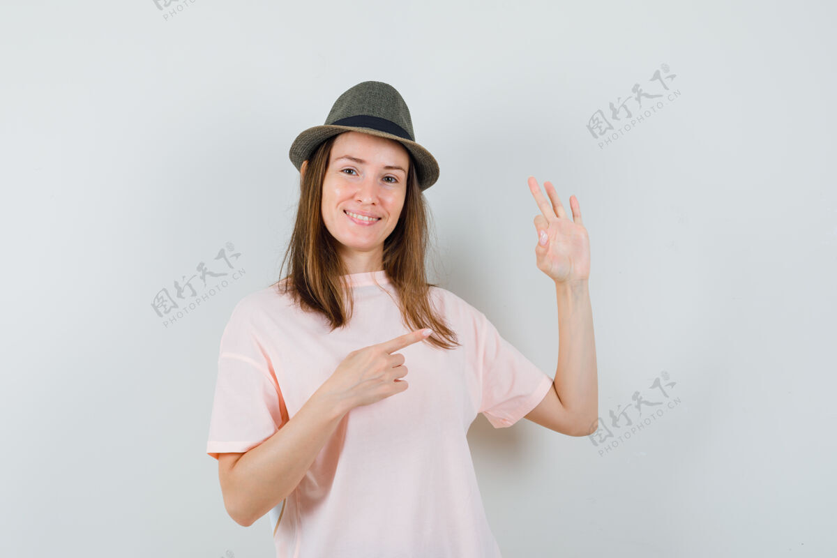 年轻年轻的女士指着旁边 展示了一个好的姿态 穿着粉色t恤 戴着帽子 看起来很自信 正面视图时尚正面Ok