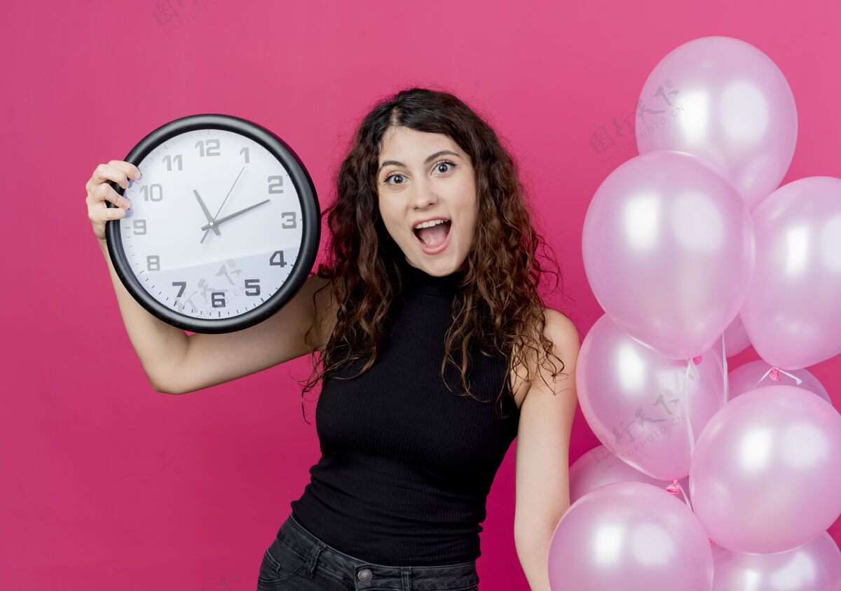 束一个年轻漂亮的女人 卷发 手里拿着一束气球 墙上挂着挂钟 站在粉红色的墙上开心地笑着气球站着抱着