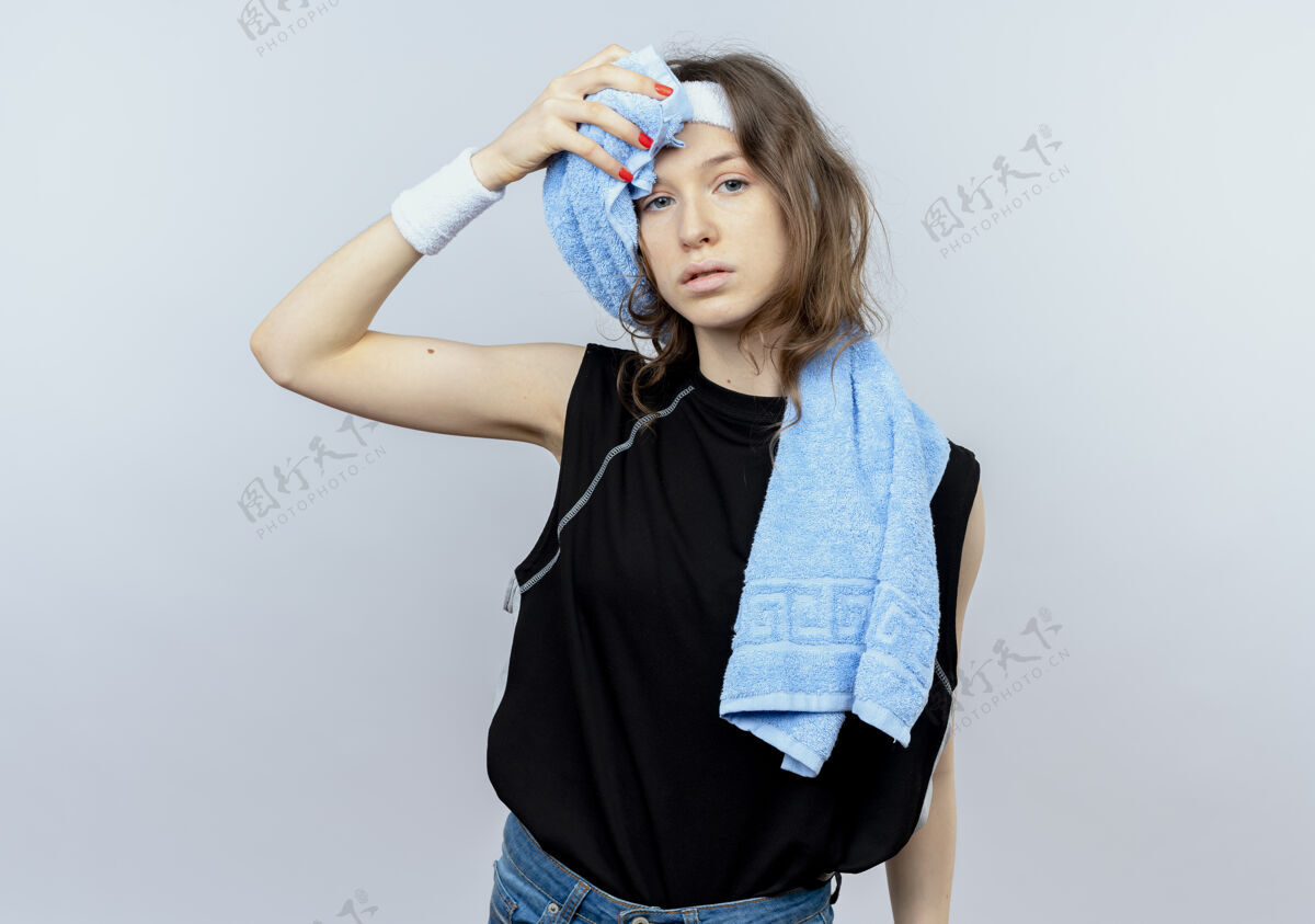 头带穿着黑色运动服 戴着头带 戴着耳机 肩上搭着毛巾的年轻健身女孩站在白墙上擦着额头 看上去很累湿巾健身耳机